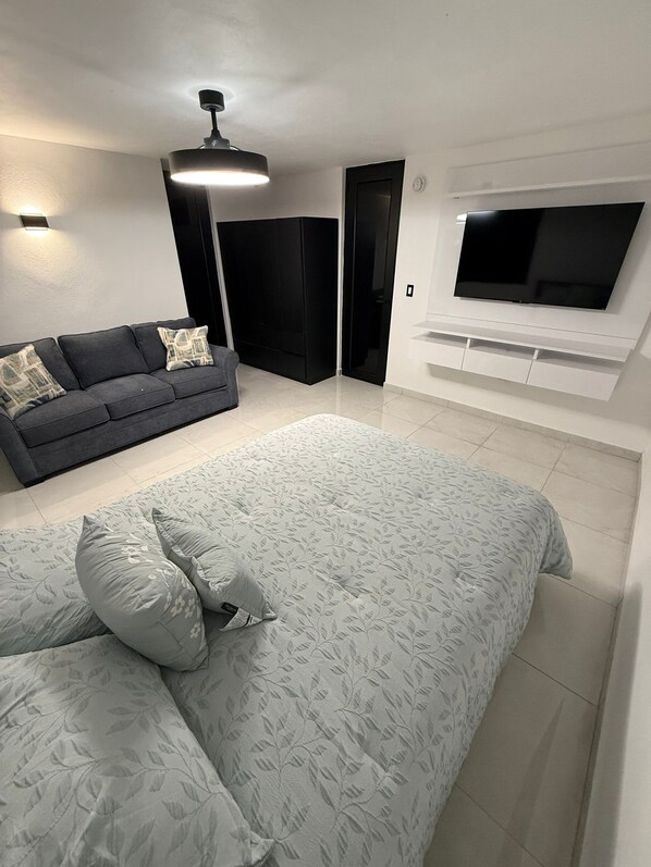 A modern bedroom with a large bed, a grey sofa, a TV mounted on the wall, and a ceiling fan.