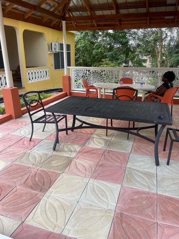 An outdoor patio with a black metal table and orange chairs, surrounded by a patterned floor and a yellow building with an air conditioning unit.