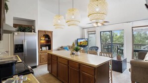 Una cucina moderna con isola centrale, mobili in legno e frigorifero integrato. Tre lampade a sospensione, un tavolo con sedie e un'ampia finestra con vista sull'esterno.