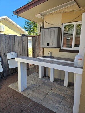 Une terrasse avec une table et des chaises blanches, un évier encastré et une fenêtre avec des stores.