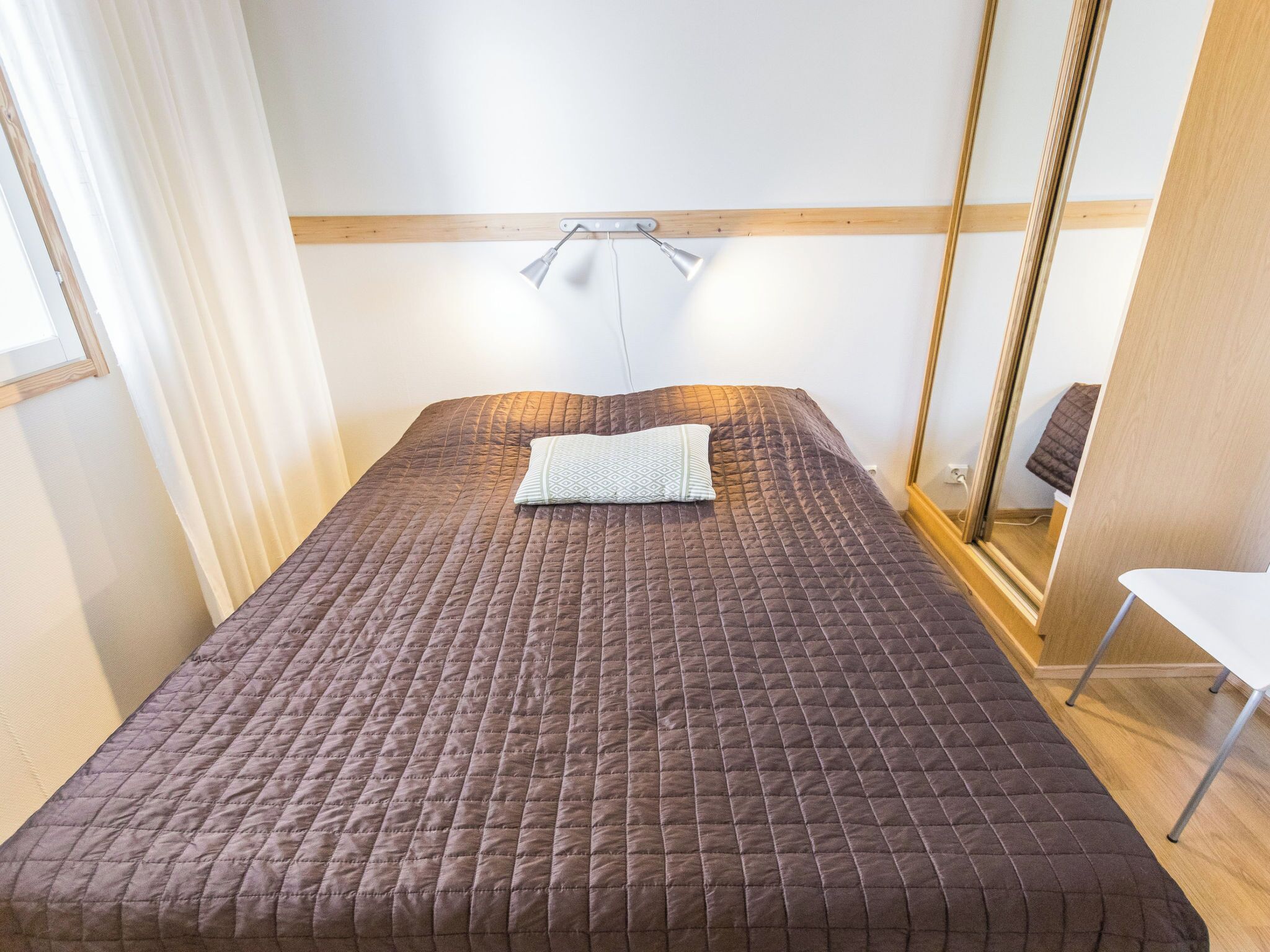 Een netjes opgemaakt bed met een gewatteerde bruine spreidek, een bedlampje en een spiegel aan de muur.