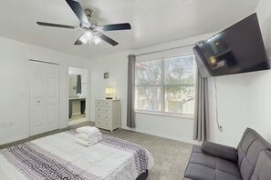 Ein Schlafzimmer mit einem Bett, einem Deckenventilator, einem an der Wand montierten Fernseher, einem Schminktisch und einem Fenster mit Vorhängen.