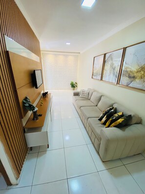 A modern living room with a sofa, a coffee table, and a TV mounted on a wooden panel. There are framed artworks on the wall and a plant on the coffee table.