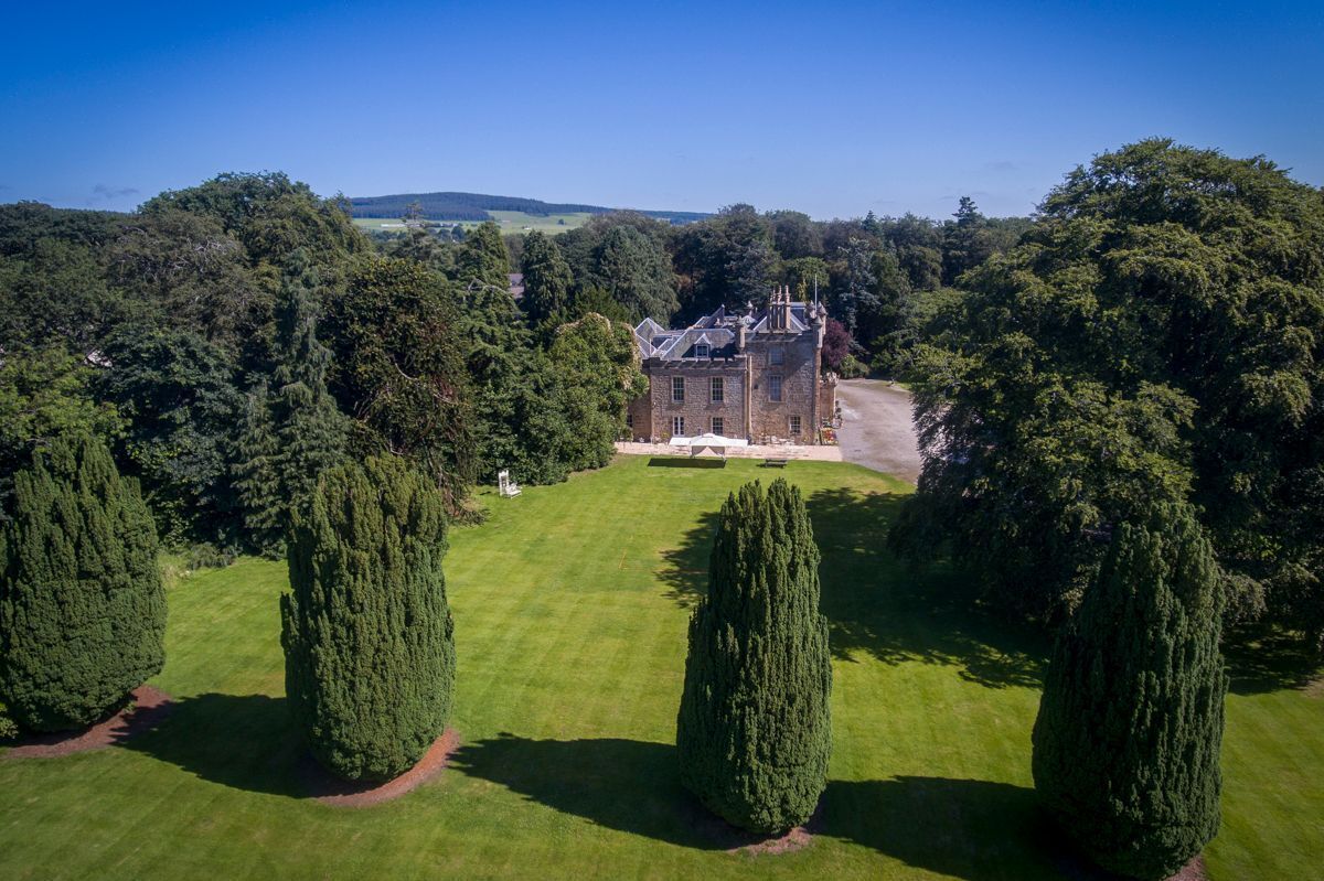 Un grand bâtiment historique en pierre, doté d’une façade symétrique, entouré de pelouses bien entretenues et de grands arbres à la forme conique.