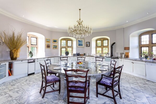A spacious dining area with a large round table surrounded by chairs, a chandelier, and a view of the kitchen through arched windows. - C17th Castle - Eight Bedroom Castle, Sleeps 16 (Clevedon)