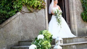 Una novia con vestido de novia blanco parada sobre escalones de piedra con flores blancas y faroles.