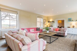 A living room with two sofas, a coffee table, and a rug. There are windows, a bookshelf, and a door leading to another room. - Emley Woodhouse - Seven Bedroom Villa, Sleeps 17 (Emley)