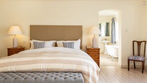 A neatly arranged bedroom with a large bed, two bedside tables with lamps, a chair, a vanity with a mirror, and a bathtub. - Snow Goose - Five Bedroom Villa, Sleeps 9 (Nutbourne)