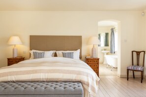 Un dormitorio ordenado con una cama grande, dos mesitas de noche con lámparas, una silla, un tocador con espejo y una bañera.