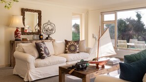 A cozy living room with a white sofa, decorative pillows, a wooden coffee table with a model sailboat, a lamp, and a mirror on the wall. - Snow Goose - Five Bedroom Villa, Sleeps 9 (Nutbourne)