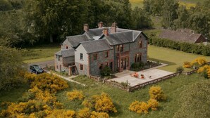 A large stone house with multiple chimneys, surrounded by a well-maintained lawn and trees.