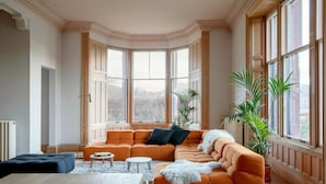 A modern living room with a large orange sectional sofa, a wooden coffee table, and a dining table. There are large windows with curtains, and potted plants are placed around the room.