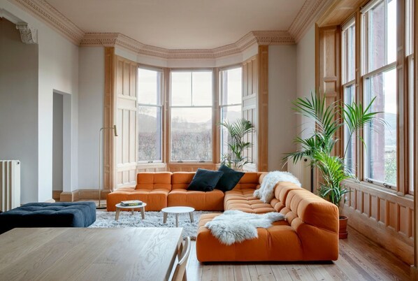 Una sala moderna con un sofá seccional naranja grande, una mesa de centro de madera y una mesa de comedor. Hay ventanas amplias con cortinas, y plantas en macetas colocadas por toda la habitación.