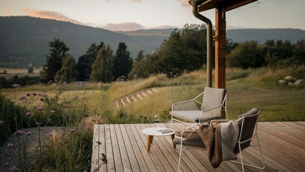 Una terraza de madera con dos sillas y una mesa pequeña, con vista a un paisaje de hierba y montañas al fondo.