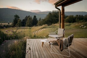 Une terrasse en bois avec deux chaises et une petite table, d’où l’on peut admirer un paysage verdoyant avec des montagnes en arrière-plan.