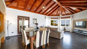 A spacious living area with a high wooden ceiling, a dining table with chairs, a sofa, a television, and a view of the outdoors. - Villa Princesa - Four Bedroom Villa, Sleeps 8 (Puerto Calero)