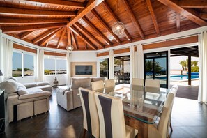 A spacious living area with a high wooden ceiling, a glass dining table, and a view of a swimming pool. - Villa Princesa - Four Bedroom Villa, Sleeps 8 (Puerto Calero)