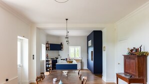 A spacious dining area with a long wooden table and chairs, a kitchen with dark blue cabinets, and a chandelier. - Maison des Grands Crus (Puligny-Montrachet)