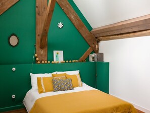 A room with a bed, a green wall, wooden beams, a framed picture, and a small shelf with books and a lamp.