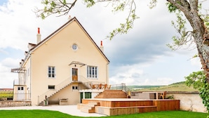 A two-story house with a wooden deck, a staircase, and a view of the countryside. - Maison des Grands Crus (Puligny-Montrachet)