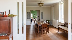 A spacious dining area with a long wooden table and chairs, a chandelier, and a view of a green room through an open door. - Maison des Grands Crus (Puligny-Montrachet)