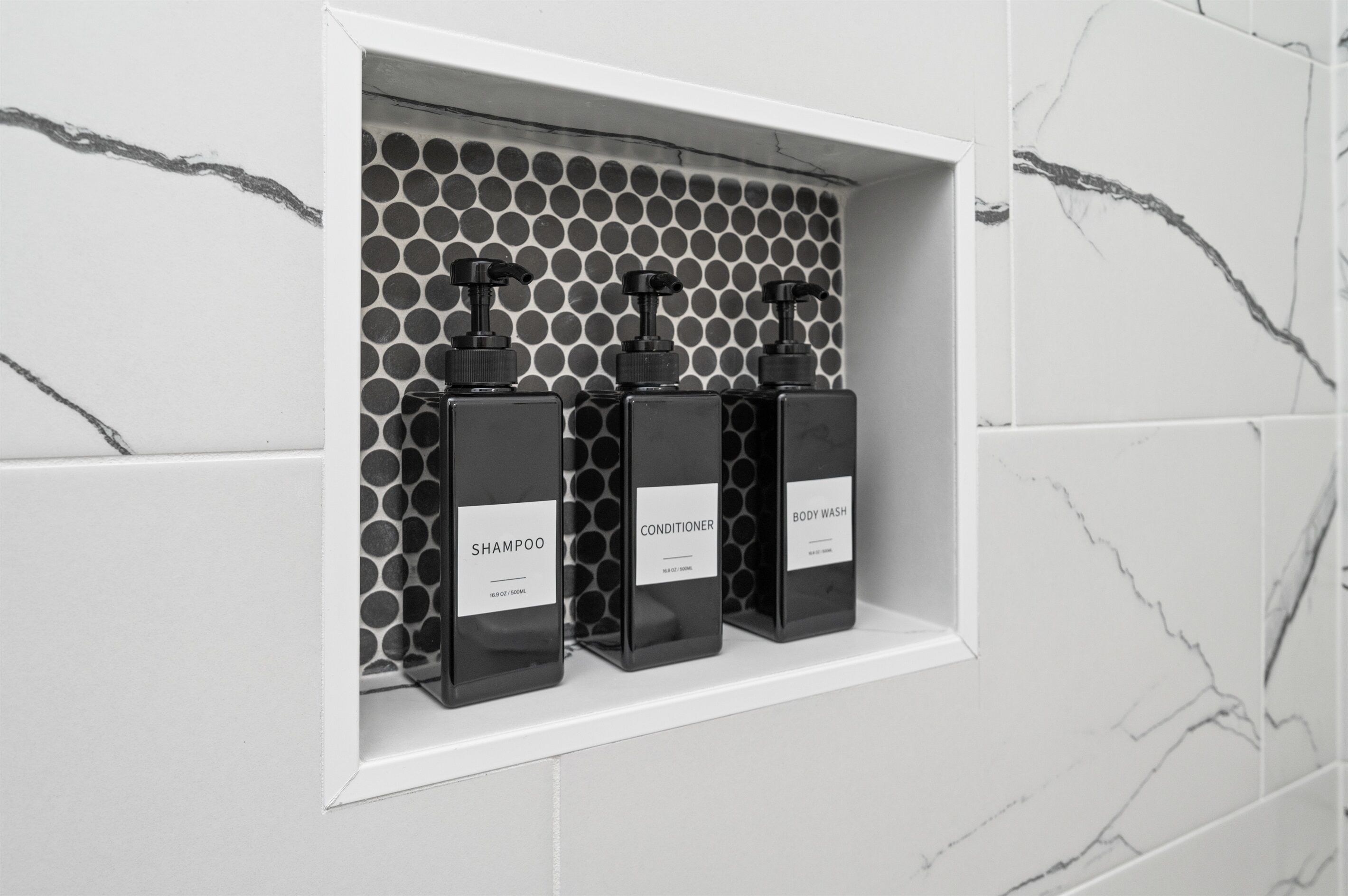 Estante de baño con tres botellas negras etiquetadas champú, acondicionador y jabón corporal.