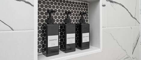 Estante de baño con tres botellas negras etiquetadas champú, acondicionador y jabón corporal.
