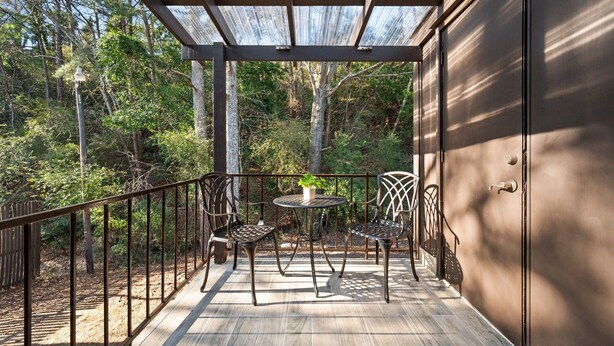 Una terraza de madera con sillas y una mesa de metal, rodeada por una barandilla metálica y un cerramiento de vidrio.