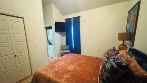 Habitación de hotel con cama, una silla, televisor, lámpara y un cuadro en la pared.