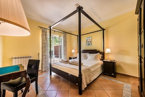 A four-poster bed with a headboard, two bedside tables with lamps, a chair, and a view of the outdoors through a window. - Villa Palmeira - Four Bedroom Villa, Sleeps 8 (Carvoeiro)