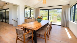 Un comedor amplio con una mesa de madera y sillas de mimbre, ventanas grandes con cortinas y vistas a zonas verdes.