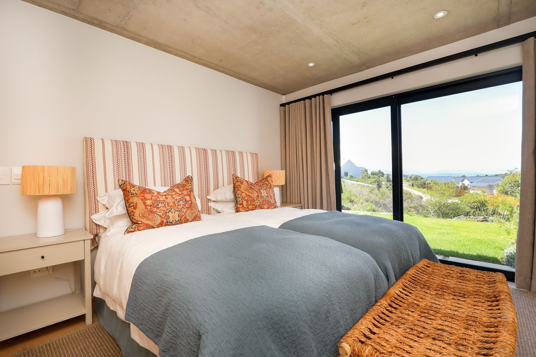 Una cama grande con almohadones estampados, una mesita de noche con lámpara, y vista a un paisaje verde a través de un ventanal amplio.