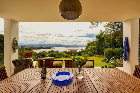 A wooden table with a view of a lake and mountains, a bottle of wine, and a blue bowl. - Busisiwe House - Four Bedroom Apartment, Sleeps 10 (Plettenberg Bay)