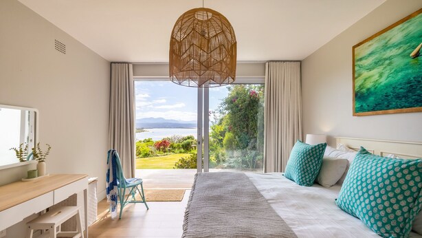 A bedroom with a large bed, a desk, and a view of a garden and water body. - Busisiwe House - Four Bedroom Apartment, Sleeps 10 (Plettenberg Bay)