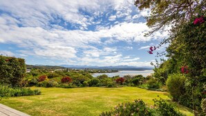 A landscaped garden with a wooden deck, lush greenery, and a view of a lake and distant mountains. - Busisiwe House - Four Bedroom Apartment, Sleeps 10 (Plettenberg Bay)