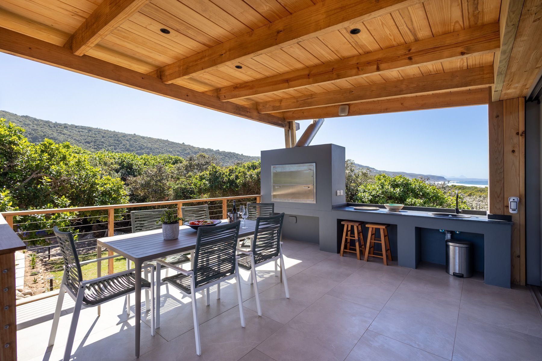 Un patio al aire libre con techo de madera, una mesa con sillas, una parrilla y una vista panorámica de colinas y el océano.