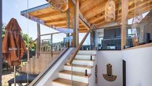 Un escalier moderne avec des marches en bois et des rampes en verre, menant à un espace extérieur doté d’une terrasse en bois et de chaises.