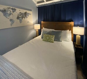 Ein ordentlich bezogenes Bett mit einer Weltkarte an der Wand, einem Kopfteil, zwei Nachttischlampen und einem blauen Vorhang.