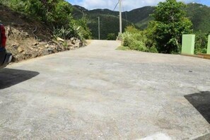 Eine gepflasterte Straße mit einem geparkten Fahrzeug links, umgeben von Grünflächen und Hügeln im Hintergrund.
