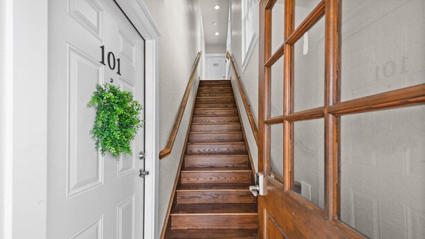 A wooden staircase with a metal handrail, white doors with numbers, and a green plant on one door. - The Abide on Magnolia (Fort Worth)