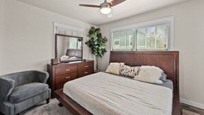 A bedroom with a bed, a dresser, a chair, a ceiling fan, and a large window. - The Abide on Magnolia (Fort Worth)