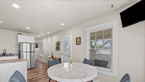A modern kitchen with a stainless steel refrigerator, a dining area with a round marble table and gray upholstered chairs, and a large window with blinds. - The Abide on Magnolia (Fort Worth)