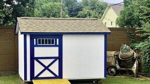 Un piccolo deposito a un piano con una porta blu e pareti bianche, una rampa in legno che conduce all'ingresso e un betoniera sullo sfondo.