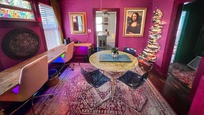 Een kamer met paarse muren, een ronde tafel met stoelen, een plafondventilator, een glas-in-loodraam en een portret aan de muur.