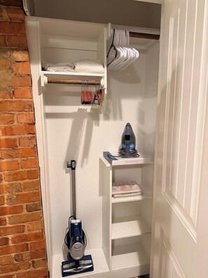 A compact storage closet with shelves, a vacuum cleaner, and a hanging rack. - Southside Lounge on Magnolia Midterm Rental (Fort Worth)