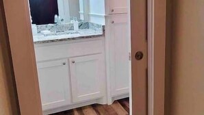 A narrow hallway leading to a bathroom with a white sink and countertop, wooden flooring, and a door with a round doorknob. - Thira on Magnolia Charming 1-bedroom house in nice Fort Worth with WiFi, AC (Fort Worth)