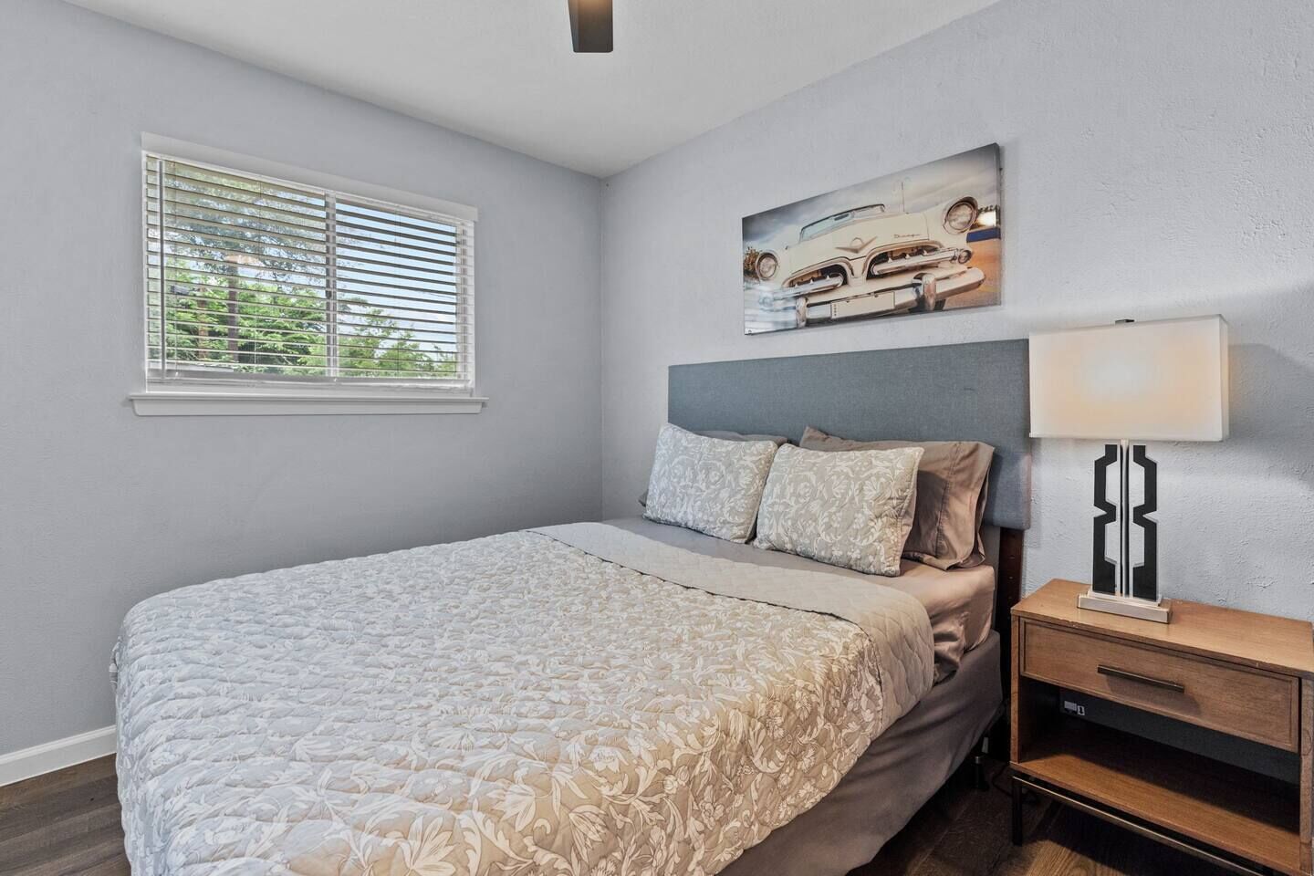 A bedroom with a bed, a nightstand, a lamp, a window with blinds, and a painting on the wall.