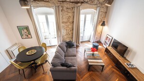 Ein modernes Wohnzimmer mit grauem Sofa, einem Holztisch und einer TV-Konsole. Es gibt eine Essecke mit einem runden Tisch und Stühlen. Der Raum verfügt über eine Steinwand und große Fenster.
