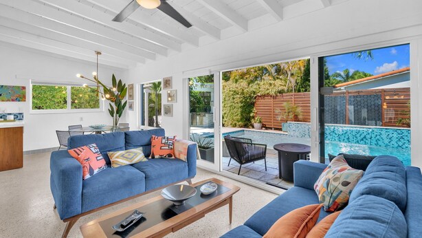 Amplia sala de estar con un conjunto de sofá azul, una mesa de centro y una mesa de comedor con sillas. Un gran ventanal corredera da a un patio exterior con piscina y plantas tropicales.