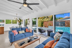 Ein geräumiger Wohnbereich mit blauem Sofa, Couchtisch und Esstisch mit Stühlen. Eine große Schiebetür öffnet sich zu einer Außenpatio mit Pool und tropischen Pflanzen.